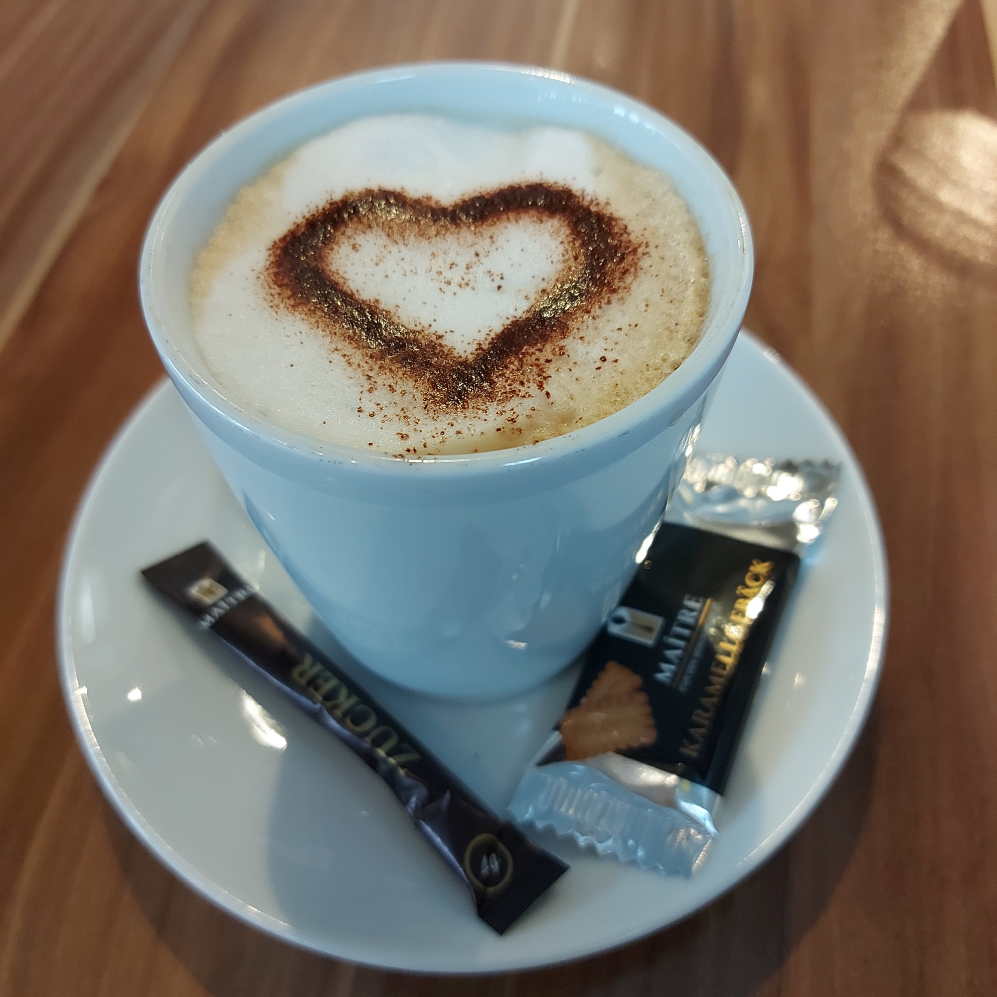 Capuccino mit einem Kakao-Herz auf der Milchschaumkrone. Auf dem weißen Untersetzer links ein Zucker und rechts ein Keks.