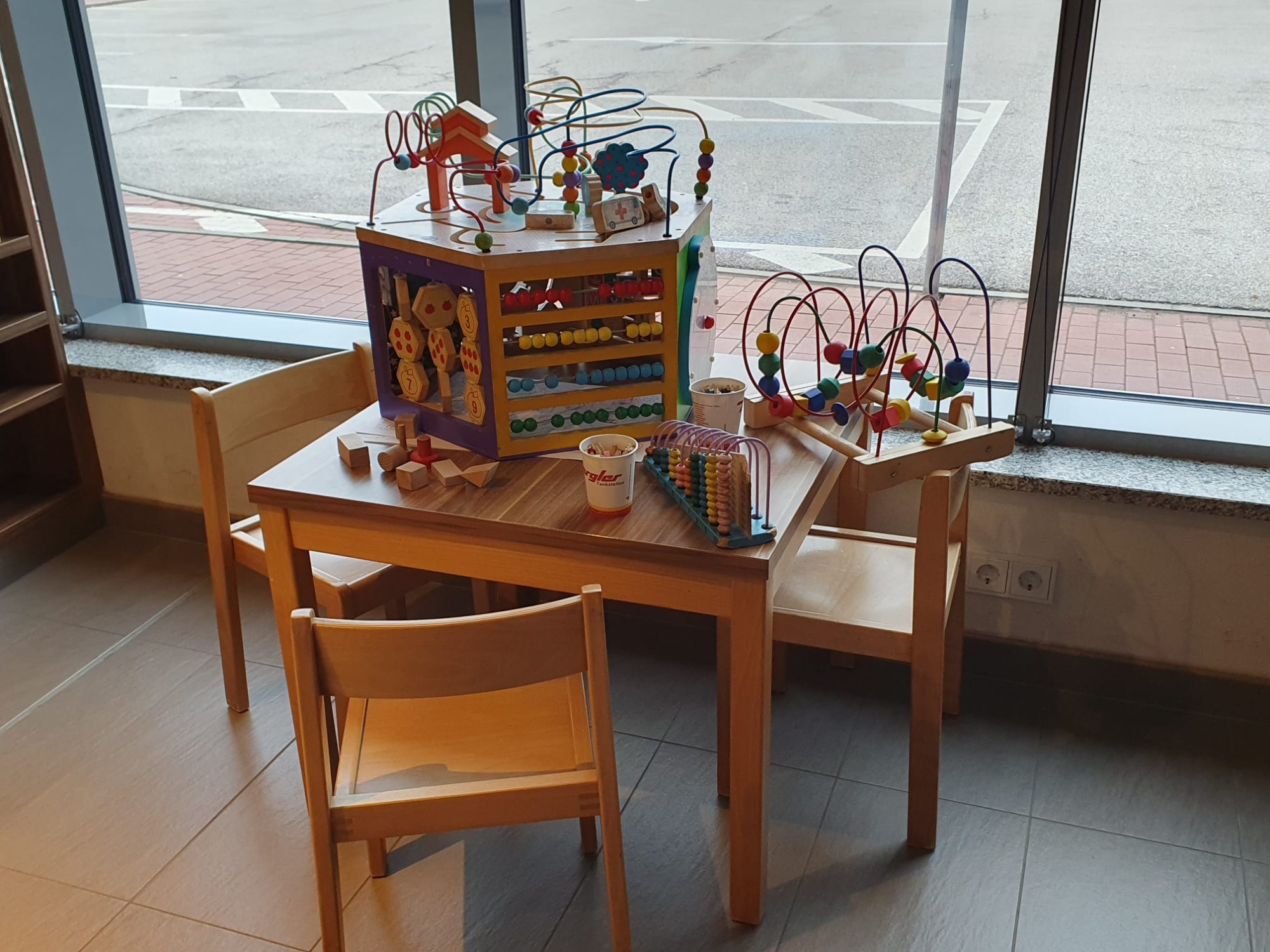 Drei Kinderstühle stehen um einen Tisch, auf dem Stifte und Motorikspiele für Kinder liegen. Aufgestellt vor einer hellen Glasfront.