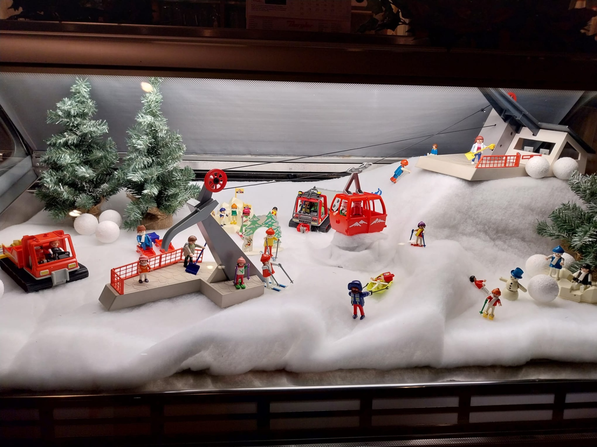 Winterlandschaft aus Playmobilfiguren. Links im Hintergrund zwei beschneite Nadelbäume, davor eine rote Pistenraupe. Im rechten Drittel ein Skilift mit Skifahrern und Rodlern.