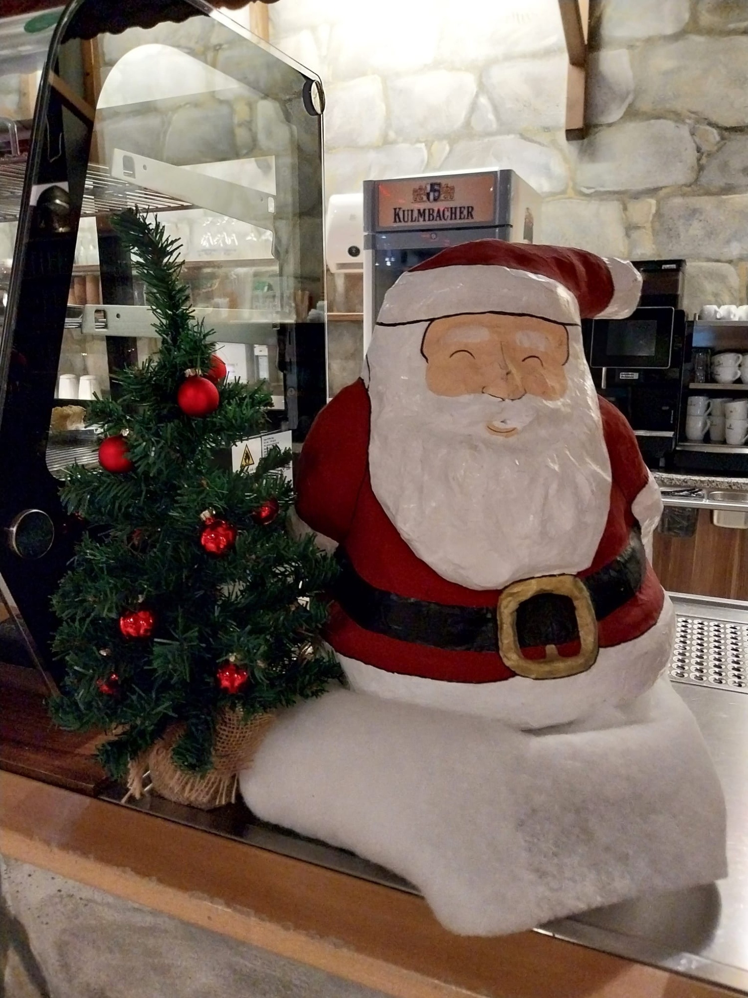 Links ein kleiner Christbaum mit roten Kugeln, daneben grinsend der Weihnachtsmann mit weißem Bart, roter Mütze und rotem Gewand.