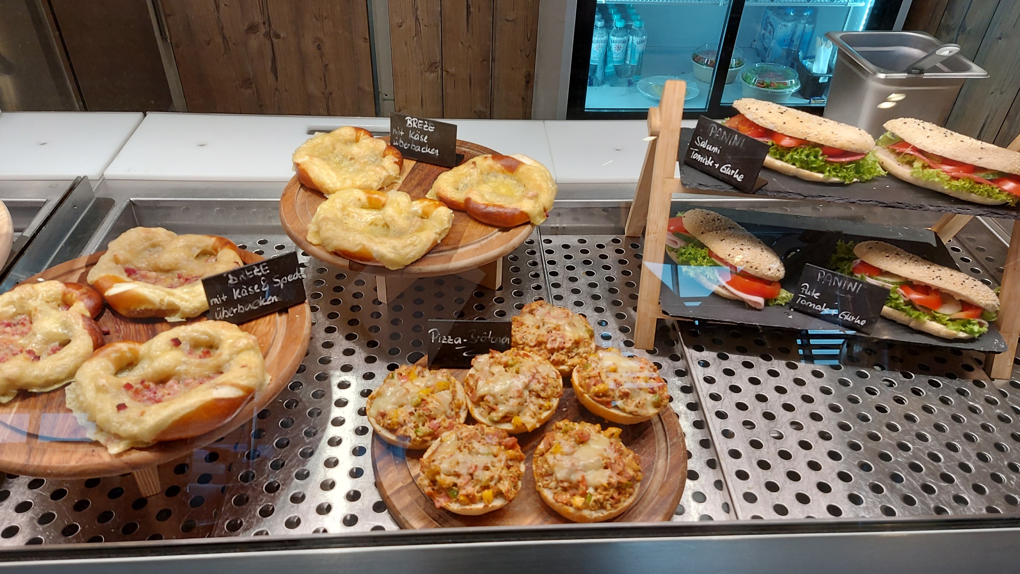 Die Theke der Brotzeithütte am Autohof. Links drei Brezen überbacken mit Speck und Käse, außerdem Pizzasemmeln jeweils angerichtet auf einem Schneidebrett aus Holz. Rechts ein Etagere mit belegten Semmeln (vegetarisch)..