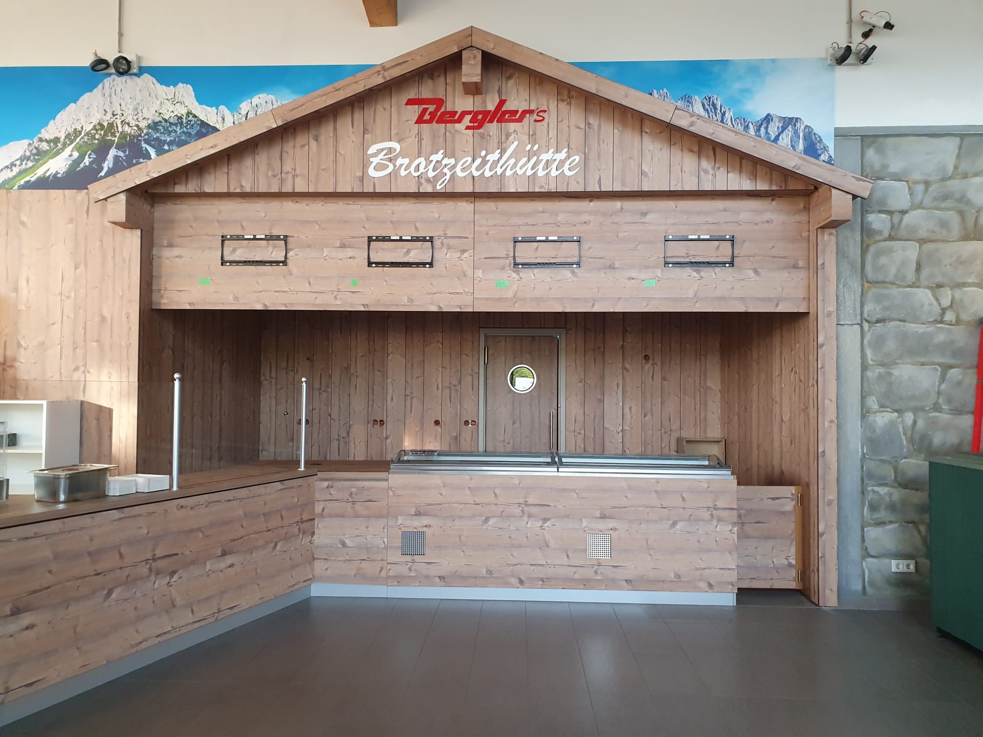 Theke in Optik einer Berghütte mit braunen Brettern, oben auf dem Giebel der Schriftzug Bergler´s Brotzeithütte. Im Hintergrund Alpenpanorama.