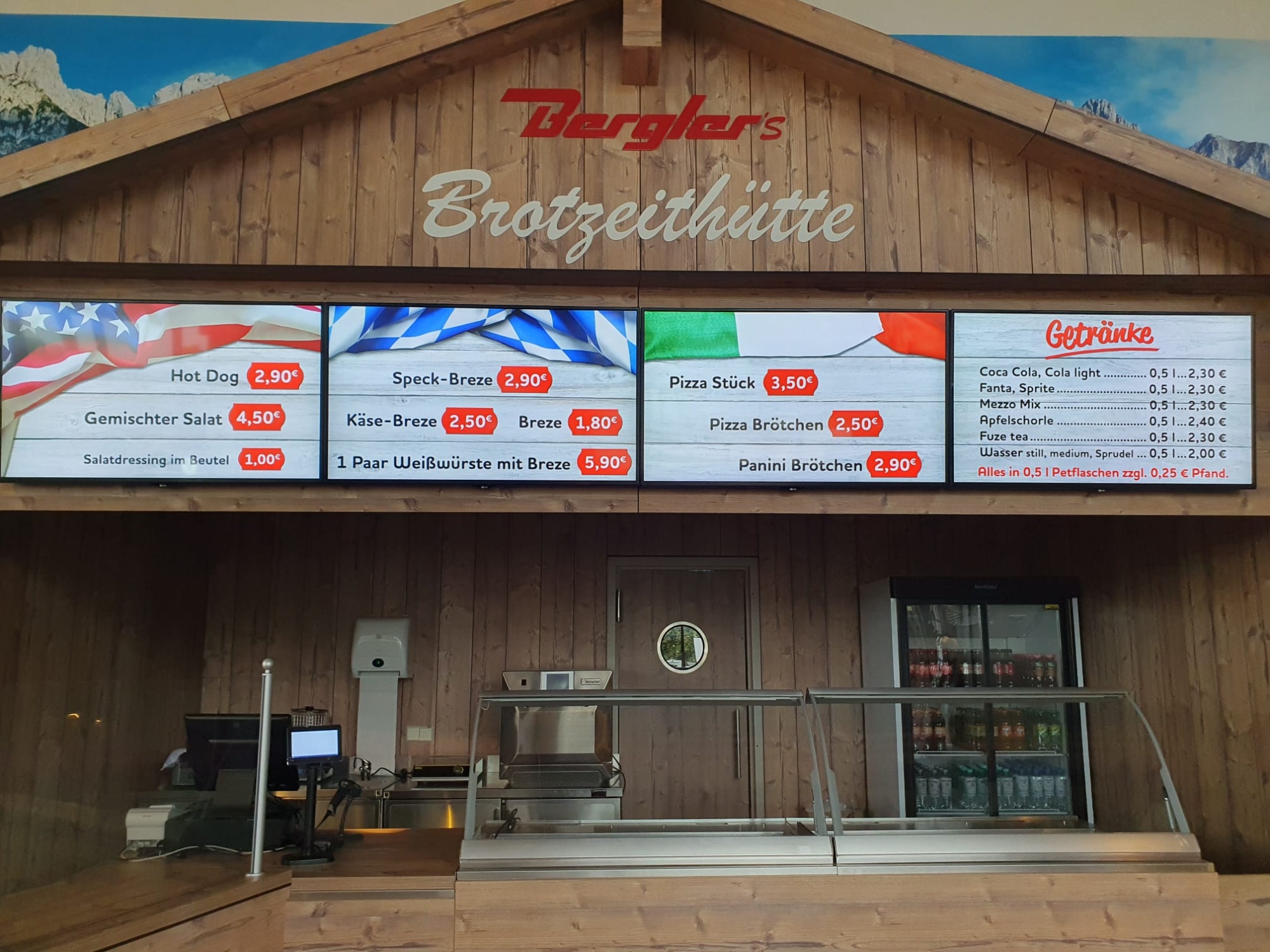 Theke in Optik einer Berghütte mit braunen Brettern, oben auf dem Giebel der Schriftzug Bergler´s Brotzeithütte. Im Hintergrund Alpenpanorama.