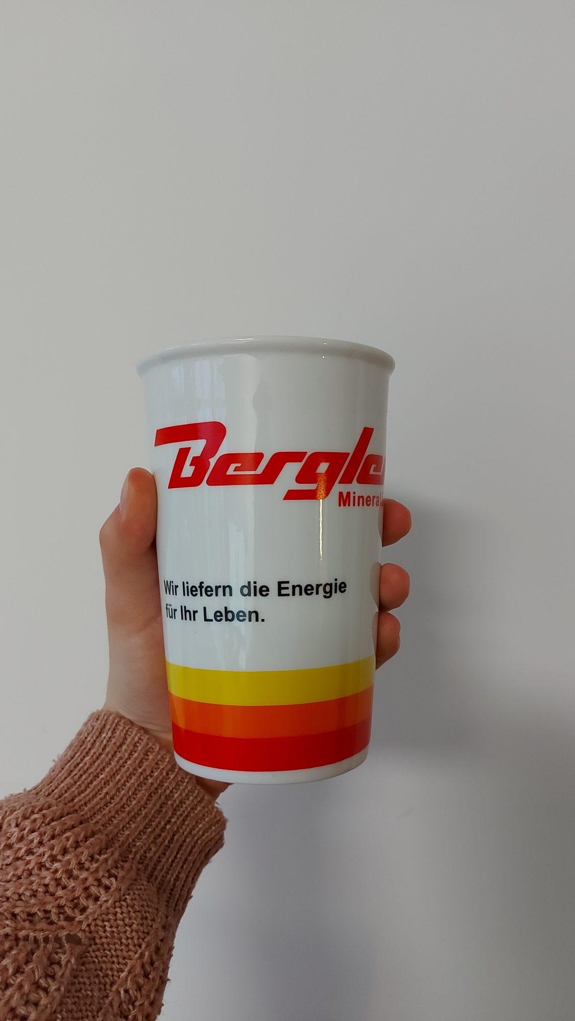 Doppwelwandiger Porzellanbecher für Kaffee wird von einer Hand gehalten. Auf dem weißen Porzellan ist in rot Bergler zu lesen. Unten wird der Becher von drei Streifen in gelb, orange und rot umrandet.