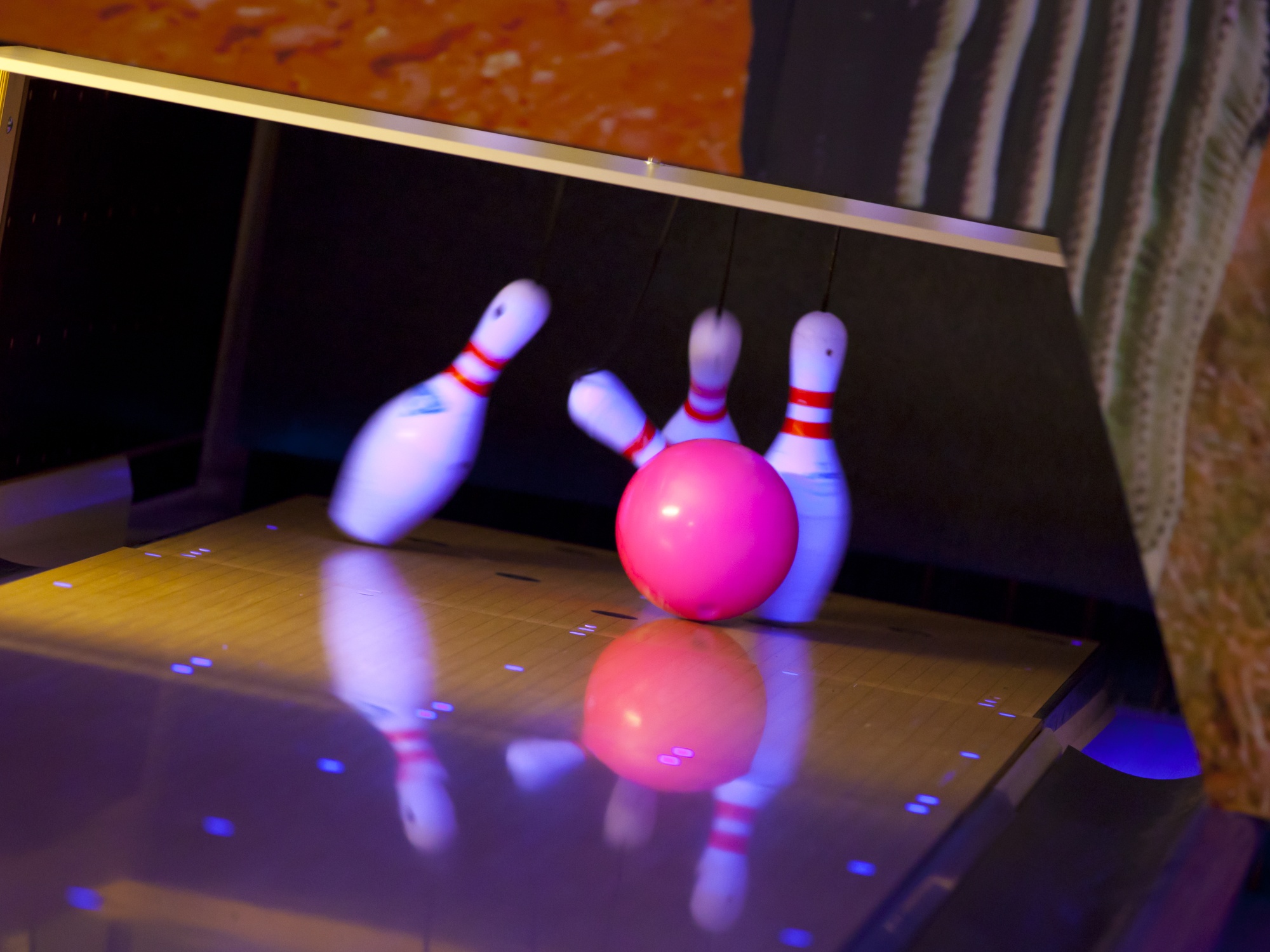 Vier weiße Bowling Pins mit zwei roten Streifen werden von einer roten Bowling Kugel getroffen und sind in der Momentaufnahme gerade am Fallen.