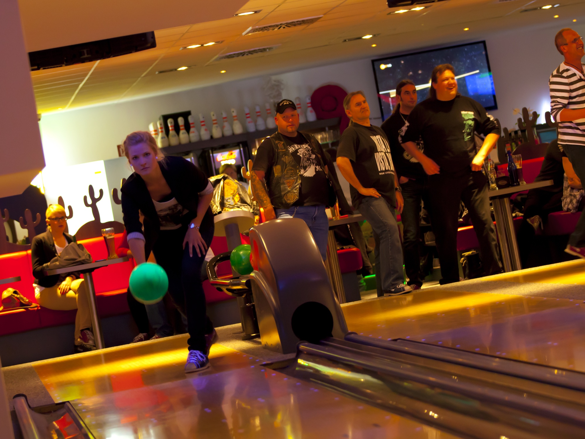 Eine Frau ist gerade dabei die Bowlingkugel Richtung Bowling-Pins zu werfen. Rechts im Bild weitere Personen, die an den anderen Bahnen spielen.